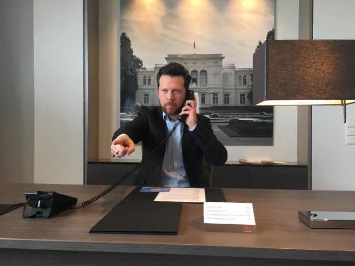 Johannes sitzt mit Jacket und Hemd an einem Schreibtisch, hat einen Telefonhörer in der Hand und zeigt mit dem anderen in das Bild, Boss-Style.