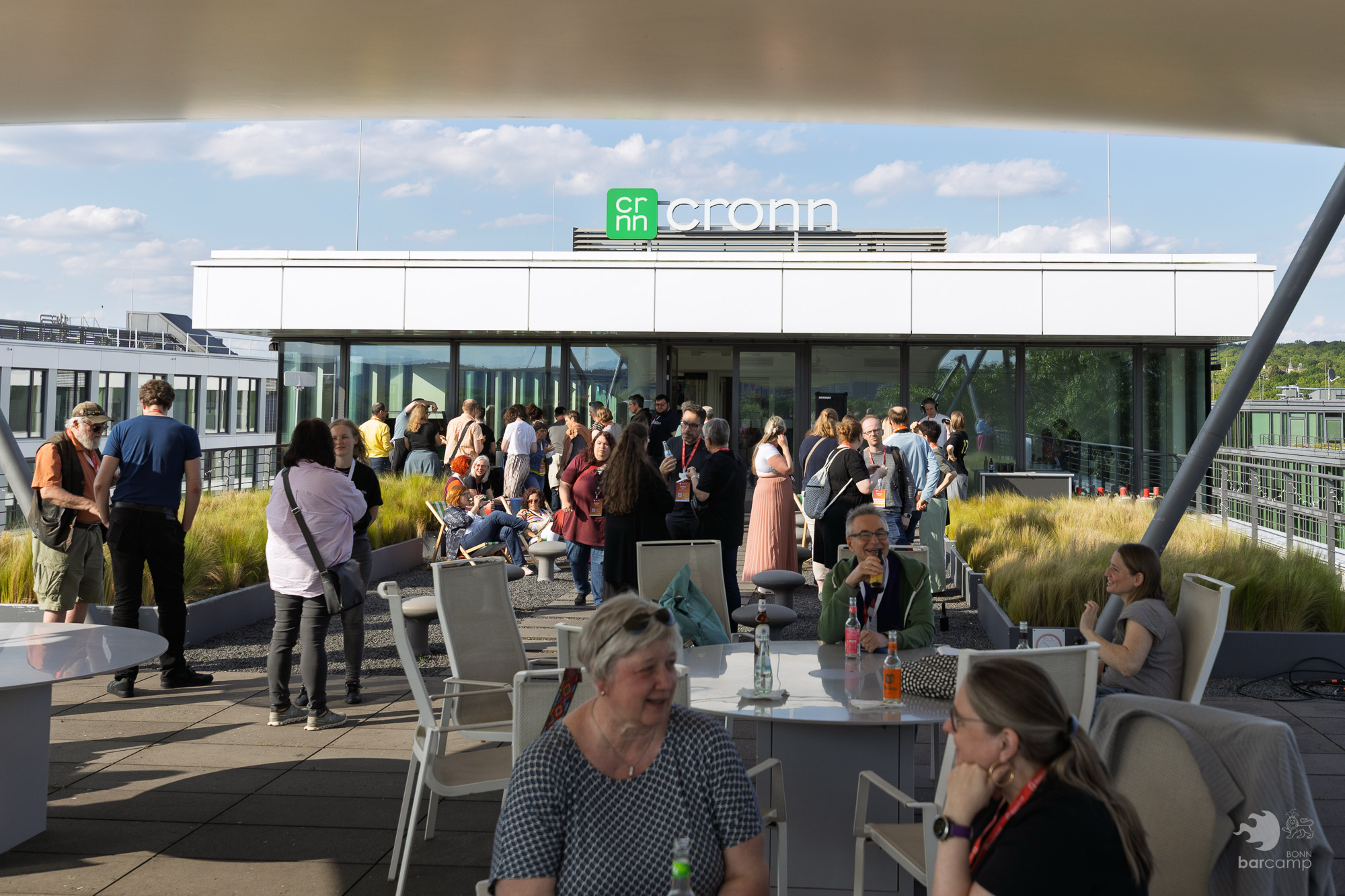 Menschen auf einer Dachterrasse mit Tischen und Stühlen vor einem Gebäude mit dem Schriftzug 'cronn'